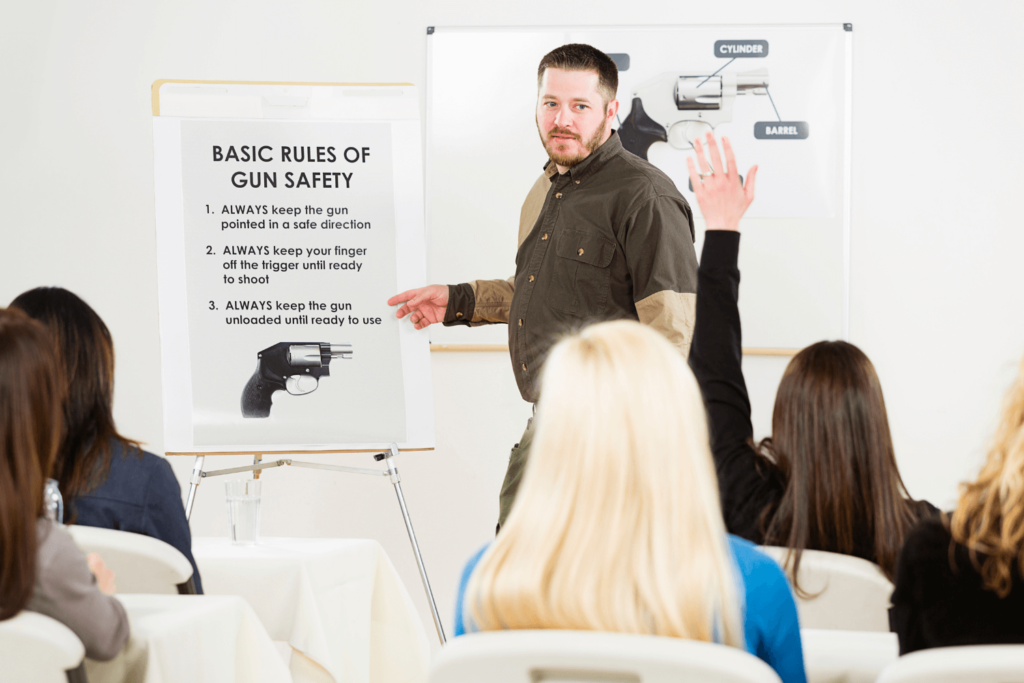 Man instructing a concealed carry class. A poster showing the basics of gun safety, and a woman in the class raising her hand