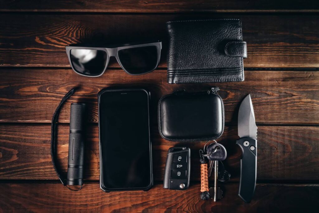 a pair of black sunglasses, wallet, phone, keys, and everyday carry knife are placed on a wooden background