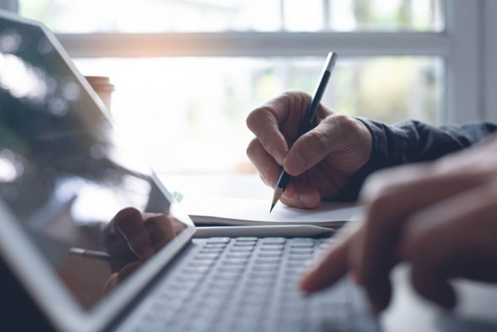 Person at a Laptop Doing Research Writing on a Notepad Next to the Laptop