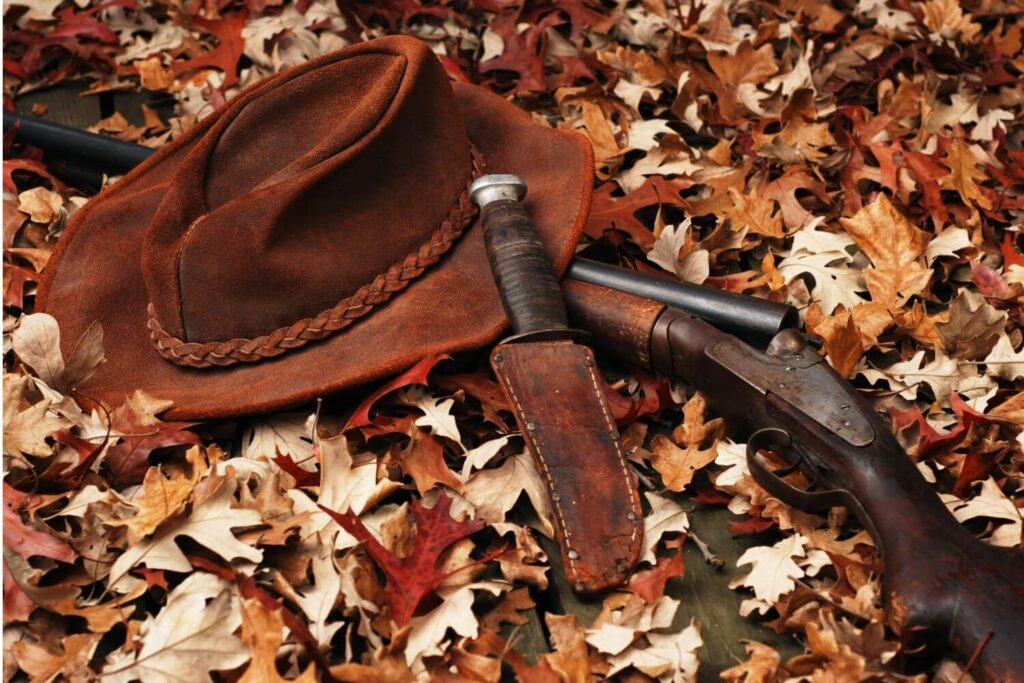 a Brown Hunter's hat, rifle, and knife in holster laying on a bed of leaves