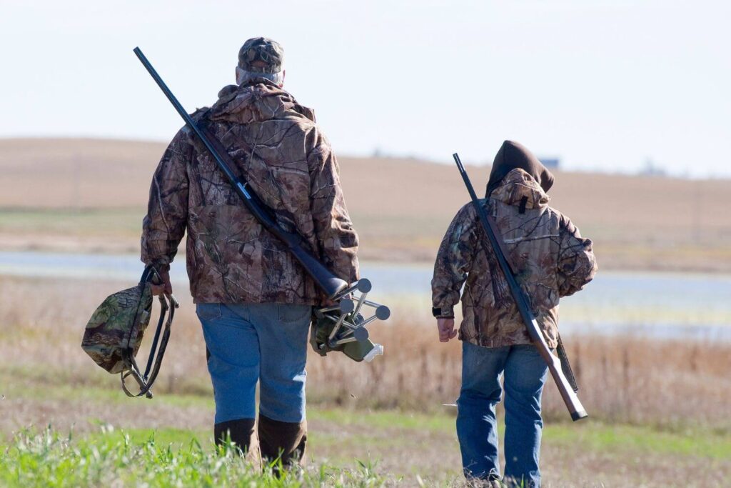 Two Hunters Dressed in Camouflage Jackets Wearing Rifles and Carrying Hunting Equipment