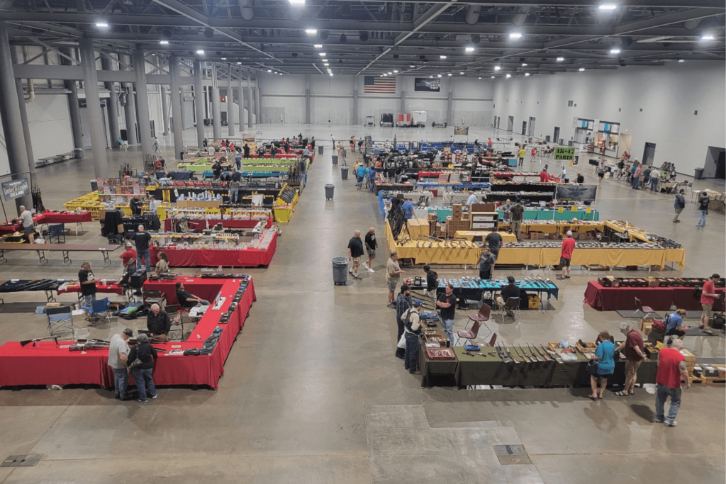Large Warehouse with Multiple Rows of Tables Set Up People Walking Around Looking at Guns and Items Laid out on Tables